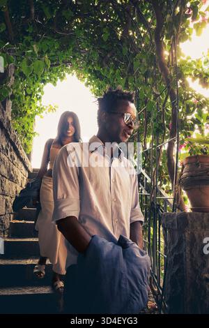 Ein charmantes Paar geht Hand in Hand entlang einer Steintreppe, die von üppigen grünen Weinstöcken beschattet wird. Die Sonne filtert durch die Blätter und erzeugt ein warmes und römisches Licht Stockfoto
