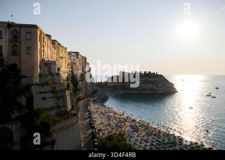 Blick aus der Vogelperspektive auf goldenes Sonnenlicht, das die alten Gebäude auf zerklüfteten Klippen küsst, im Kontrast zum türkisfarbenen Meer und dem geschäftigen Strand darunter. Stockfoto