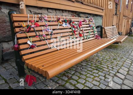 Eine lange Holzbank steht auf einem Kopfsteinpflasterweg neben einem rustikalen Gebäude. Farbenfrohe Strickgirlanden und Blumen bedecken die Rückenlehne und schaffen einen charmanten Look Stockfoto