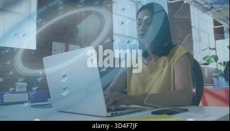 Reife Frau mit silbernem Laptop im Büro mit Grafik-Overlay, Senfoberteil Stockfoto