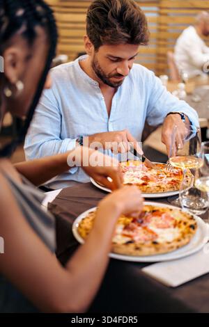Zwei Freunde sitzen an einem Tisch und teilen sich eine leckere Pizza. Der Mann konzentriert sich darauf, seine Scheibe zu schneiden, während die Frau sich darauf vorbereitet, ihre zu genießen. Das Ambiente ist W Stockfoto