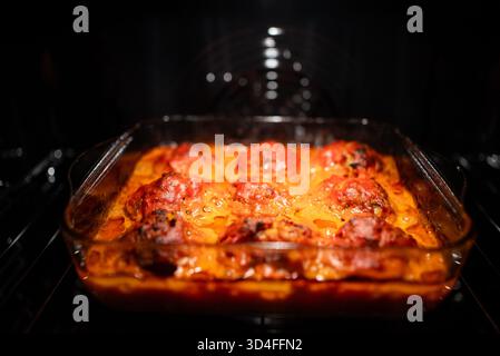 Fleischbällchen, die in reichhaltiger Tomatensauce in einer Auflaufform aus Glas köcheln, im dunklen Ofen braten – warme, hausgemachte Komfortgerichte für ein Familienessen Stockfoto