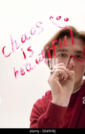 Ein junger Mann in modischer Kleidung schreibt kreativ auf einer transparenten Oberfläche und zeigt seinen Stil. Stockfoto