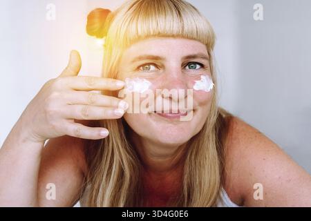 Attraktive Blondine mit blauäugigen Augen mittleren Alters, mit gebundenen Pferdeschwänzen auf den Haaren, sieht im Badezimmer im Spiegel, trägt Creme gegen Falten, Altersflecken auf, wh Stockfoto