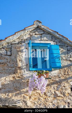 Bezauberndes, sonnendurchflutetes Steinhaus mit leuchtend blauen Fensterläden, einem Fensterkasten mit weiß-violetten Blüten, rustikalen strukturierten Wänden und einem hellen Himmel, der an Mediter erinnert Stockfoto
