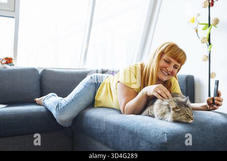 Portrait positive blonde Frau mittleren Alters, die auf dem Sofa liegt und eine Katze umarmt, die Selfie macht. Das Leben zu Hause mit einem Haustier Stockfoto