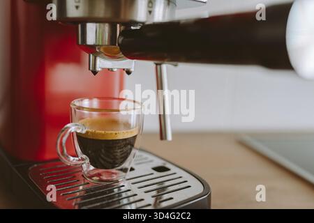 Espresso, der in der Küche in eine transparente Tasse aus der Kaffeemaschine gegossen wird, vor verschwommenem Hintergrundlicht Stockfoto