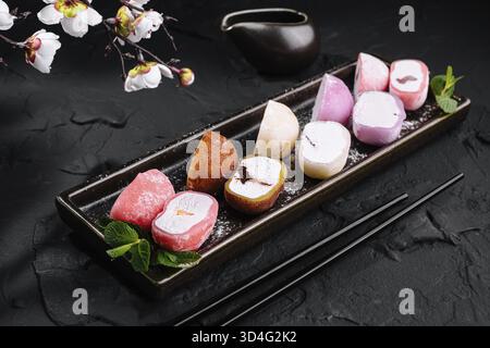 Speisen, japanische Mochi-Eissorten, serviert auf einem Keramiktablett mit Kirschblüten, dunkle Ästhetik Stockfoto