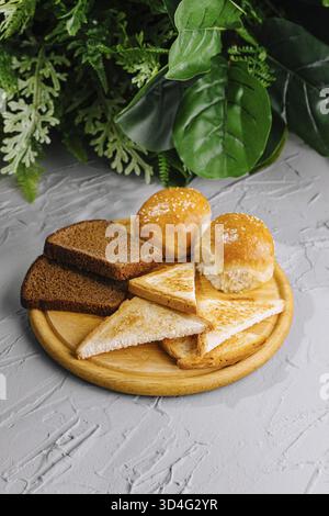 Essen, Auswahl an frisch gebackenem Brot auf einem runden Holzschneidebrett, mit grünen Pflanzen im Hintergrund Stockfoto