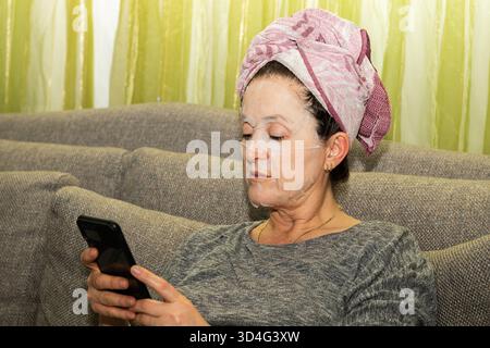 Eine Frau, die eine Papiermaske trägt, entspannt sich auf einem bequemen Sofa zu Hause mit ihrem Handy. Schönheitsroutine und -Technik. Stockfoto