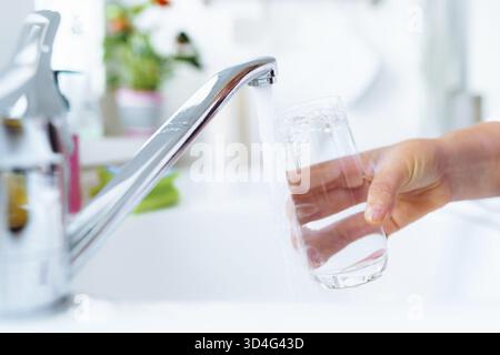 Die Hand der Frau hält ein Glas sauberes Wasser in der Nähe des Wasserhahns in der Küche. Das Streuwasser wird in das Glas gegossen Stockfoto