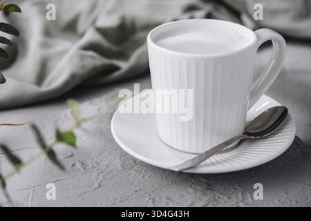Essen, genießen Sie einen gemütlichen Morgen mit einem aufschäumenden Milch-Cappuccino in einer einfachen und eleganten weißen Tasse auf einem strukturierten Tisch umgeben von grünen Blättern Stockfoto