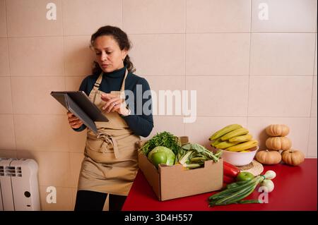 Eine Frau in einer beigefarbenen Schürze überprüft ein Tablett neben einer Schachtel mit frischem Gemüse und Obst auf einer roten Theke und hebt Lebensmitteleinkäufe, Inventar oder Onl hervor Stockfoto