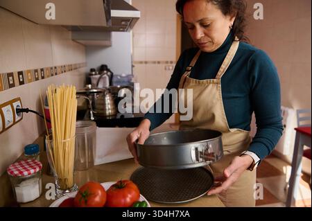 Eine Frau in einer Schürze steht an einer Küchentheke und hält eine runde Backform. Frische Tomaten, Nudeln, Gläser und Utensilien umgeben sie, während sie einen zubereitet Stockfoto