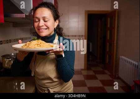 Eine fröhliche Frau in einer Schürze hält einen Teller Pasta mit Basilikum in einer warmen Küche und feiert Hausmannskost und einen gemütlichen Dinner-Moment. Stockfoto