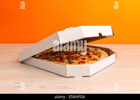 Pizza in halb geschlossener Box auf Holztisch mit orangem Hintergrund, symbolisiert frische Lieferung, Fast Food und moderne Restaurantverpackung Stockfoto