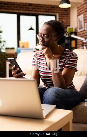 Eine nachdenkliche schwarze Frau, die mit der Hand am Kinn auf der Couch sitzt, liest ernste Nachrichten auf dem Smartphone. Nachdenkliche Freiberuflerin, die ein mobiles Gerät mit besessenem Ausdruck betrachtet, erhält unerwartete Nachrichten Stockfoto