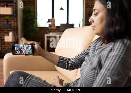 Unternehmerin im virtuellen Gespräch mit internationalen Fachleuten während der Online-Sitzung. Nahaufnahme zeigt eine inderin mit Smartphone, die aktiv an Videokonferenzen vom Sofa aus teilnimmt. Stockfoto