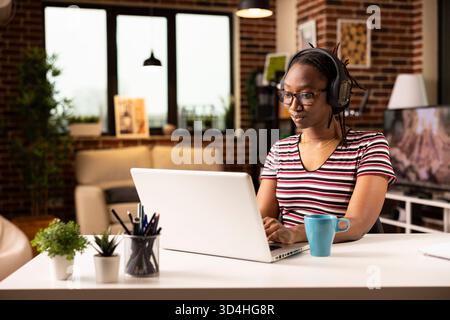 Afroamerikanerin, die Kopfhörer trägt, Online-Artikel liest und Geschäftsverträge auf Laptop überprüft. Schwarze Freelancerin genießt Musik, während sie von zu Hause aus mit Kaffee in der Nähe arbeitet. Stockfoto