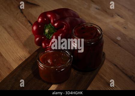 Paprika und zwei Gläser hausgemachter roter Pfeffer-Zubereitungen auf Holzoberfläche Stockfoto