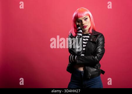 Die wunderschöne rosafarbene Frau posiert mit Hand auf Wange, trägt eine Lederjacke und ein gestreiftes Hemd und strahlt im Studio eine kühne Eleganz aus. Weibliches Modell mit lebendigem Haar und ruhigem Ausdruck posiert anmutig. Stockfoto