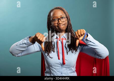 Afroamerikanerin mit den Lippen, die zwei Daumen nach unten drückt, und eine starke Missbilligung ausdrückt. Schwarze Frau mit rotem cape, in ernster Pose, die Kühnheit und Authentizität widerspiegelt. Stockfoto
