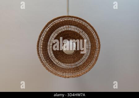 Nahaufnahme eines gewobenen Rattan-Lampenschirms mit Glühbirne, der von einer weißen Decke in Malbork hängt. Konzentrische Korbringe bilden einen warmen, minimalistischen Look Stockfoto