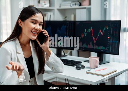 Lächelnde junge, schöne asiatische Geschäftsfrau, die in einem modernen Büro einen Broker auf dem Smartphone anruft, um Börsenkurven auf dem pc-Bildschirm zu analysieren Stockfoto