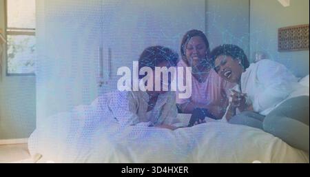 Lachende drei Frauen in Loungewear, die sich auf einem Queensize-Bett im Schlafzimmer entspannen und ihr Smartphone teilen Stockfoto