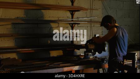 Schneidemann mit Tischsäge in schwacher Werkstatt, ärmelloses Hemd, langer Stock, Metallstäbe, Kopierraum Stockfoto