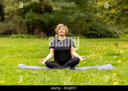 Eine ältere Frau sitzt friedlich in der Natur und übt Yoga auf einer Matte. Die Umgebung ist ruhig mit Grün und Herbstfarben. Ihre Konzentration und Ruhe Stockfoto