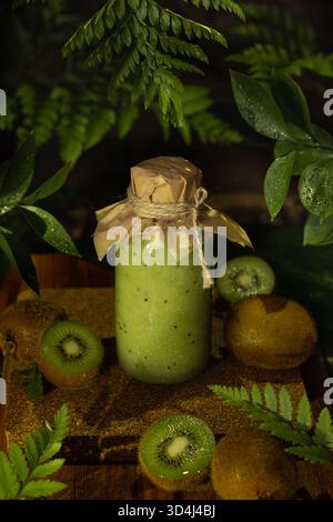 Ein erfrischender grüner Smoothie wird in ein Glas gegeben, das mit einem Papierdeckel bedeckt ist und von geschnittenen Kiwis und grünen Blättern umgeben ist, was auf eine Gesundheit hindeutet Stockfoto