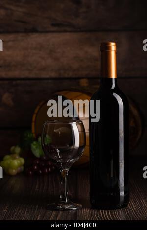 Eine Weinflasche steht hoch neben einem leeren Glas auf einem Holztisch. Trauben umgeben die Gegenstände, mit einem Fass im Hintergrund, das ein gemütliches Atmosp erzeugt Stockfoto
