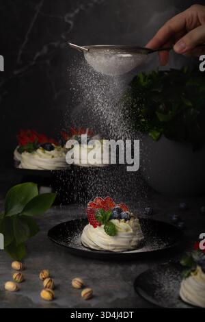 Eine Hand streut sanft Puderzucker über elegante pavlova-Desserts mit lebendigen Erdbeeren und Heidelbeeren. Üppiges Grün sorgt für Frische Stockfoto