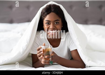 Fröhliche Frau schlürft Kräutertee im Bett und genießt ein gemütliches Erlebnis am Morgen Stockfoto