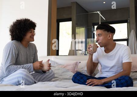 Verschiedene männliche Freunde, die im Kreuz auf dem Bett sitzen, im modernen Schlafzimmer, mit Keramikbecher Stockfoto