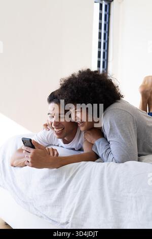 Lachende männliche Freunde, die nebeneinander im Schlafzimmer liegen und ihr Smartphone auf weißem Bett teilen Stockfoto