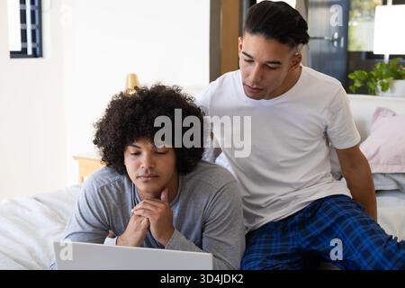 Verschiedene männliche Freunde, die sich auf dem Bett in einem hellen Schlafzimmer mit silbernem Laptop-Computer beugten Stockfoto
