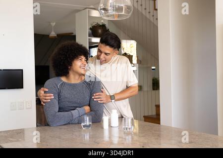 Lächelndes, vielfältiges männliches Paar, das sich an der Küchenarbeitsplatteninsel umarmt, mit Glaspendelleuchte Stockfoto