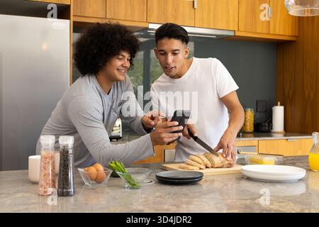 Verschiedene männliche Freunde bereiten Frühstück in moderner Küche mit Smartphone und Küchenmesser zu Stockfoto