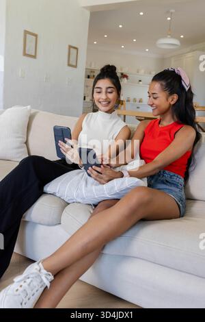 Auf Smartphones surfen verschiedene Freundinnen, die sich auf dem Sofa in einem hellen Wohnzimmer mit Kissen entspannen Stockfoto