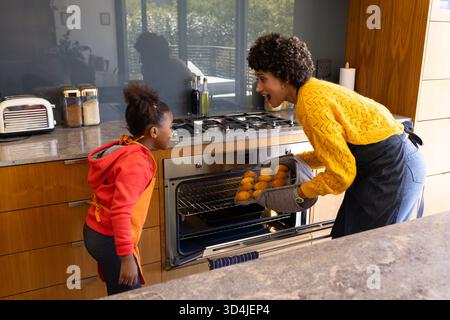 Mutter und Tochter ziehen Muffins aus dem Ofen in der modernen Küche, mit Gläsern und Toaster Stockfoto