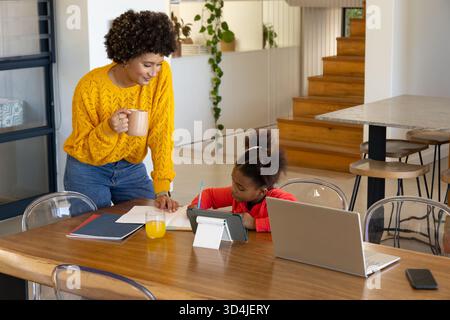 Vielseitige Mutter und Tochter, die in der Wohnküche auf dem Tisch sitzen, mit Notebook und Tablet Stockfoto