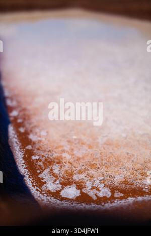Eine Nahaufnahme von weißen Meersalzkristallen, die sich auf einer rötlich-braunen Ton- oder Schmutzoberfläche bilden, möglicherweise in einem Salzdampfteich. Ein dunkelblaues Ed Stockfoto