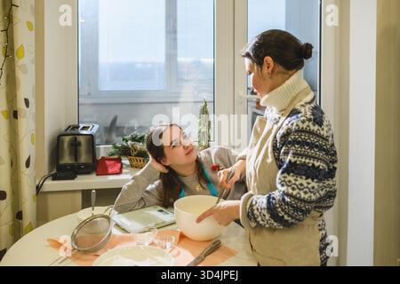 Lächelnde Frau und ihre Tochter bereiten in der gemütlichen Küche Teig für Weihnachtskuchen vor. Warmes Sonnenlicht, festliche Stimmung, Familienkochzeit, Urlaubsvorbereitungen Stockfoto