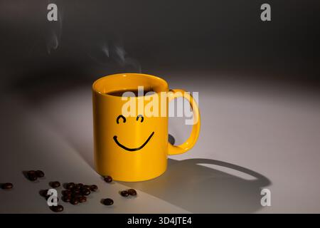 Eine gelbe Kaffeetasse mit einem Smiley-Gesicht sitzt auf einer Oberfläche, nahe einer Verstreuung von Kaffeebohnen. Dampf steigt aus dem Becher, was auf ein warmes und einladendes Getränk hindeutet. Stockfoto