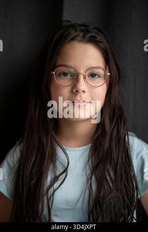 Vertikales Porträt eines Mädchens mit langen Haaren und Brillen, ernstes Gesicht vor dunklem Stoffhintergrund, weiches Licht, Kopierraum. Konzept von Fokus, Tiefe Stockfoto