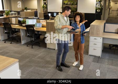 Diskussion verschiedener Kollegen, die in der Nähe einer Trennwand im Großraumbüro stehen, mit Tablet und Notebook Stockfoto
