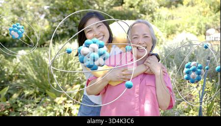 Umarmte Tochter in gelbem Oberteil, umarmte Mutter in rosa Hemd im Garten mit Metallskulpturen Stockfoto