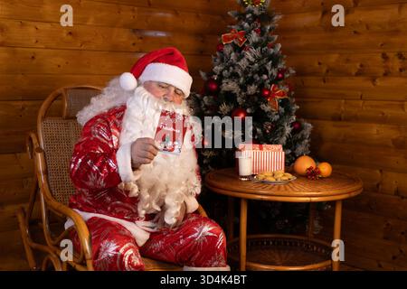 Der Weihnachtsmann hält eine Weihnachtskarte in der Hand, während er auf einem Stuhl sitzt Stockfoto